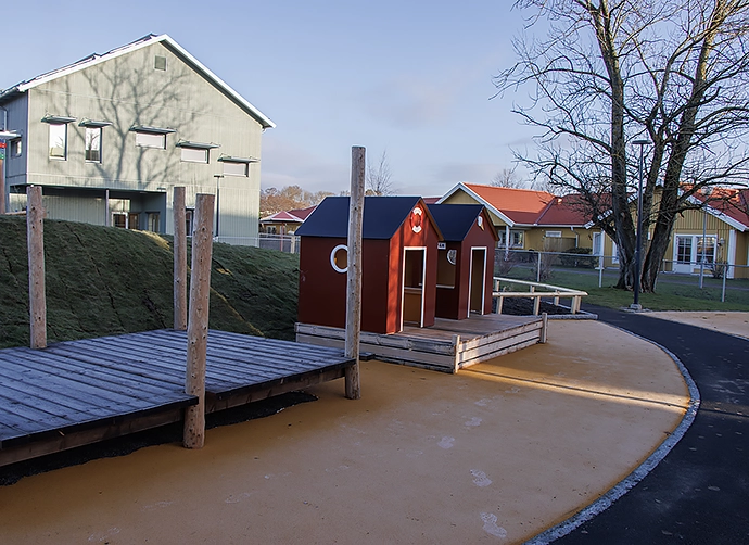 Förskolegård med röda fiskebodar och en brygga och förskolan i bakgrunden