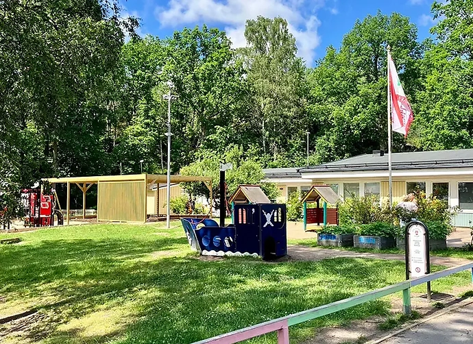 Gräsbeklädd förskolegård med båt framför förskola med höga träd bakom