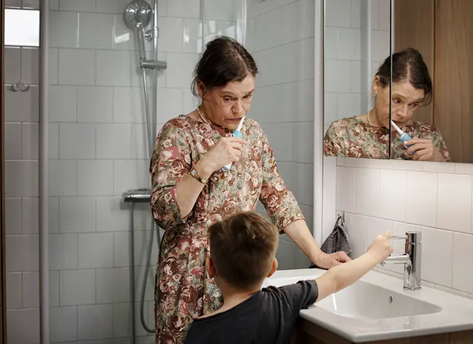 En kvinna står i ett badrum och borstar tänderna. Kvinnans barn stänger av kranen så den inte står och rinner under tiden.