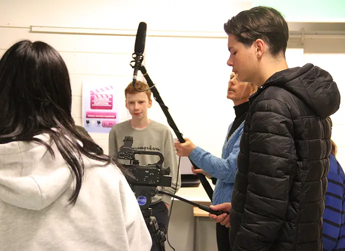 En filminspelning med fyra ungdomar och en lärare som står runt en filmkamera på ett stativ. En kille håller i stativet och läraren håller upp en ljudbom.