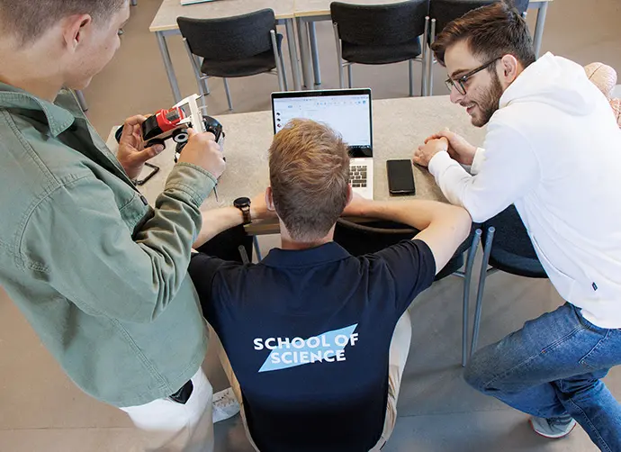 Elever arbetar tillsammans kring en dator och analyserar en teknisk modell, sett uppifrån.