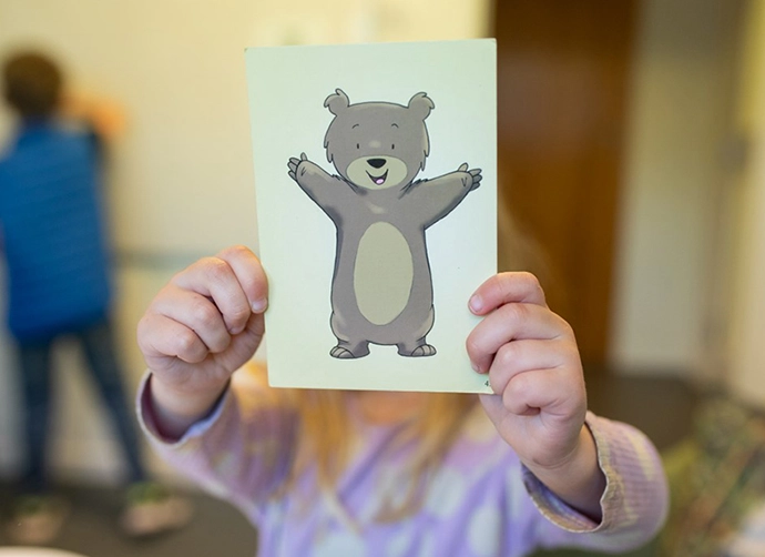 Ett barn står i mitten av bilden och håller upp ett kort framför sitt ansikte. På kortet syns en illustration av en nalle som ser glad ut och som sträcker ut armarna mot betraktaren. Fotot är taget inomhus. I bakgrunden står ett annat barn som skriver något på en whiteboard.