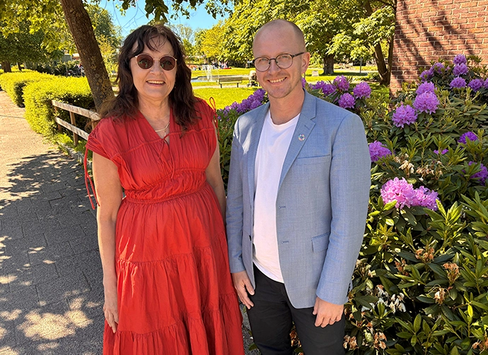 Caharina och Johan står bredvid varandra. De tittar in i kameran och ser glada ut. Till höger syns en blommande rhododendron och i bakgrunden syns stadsparken. Det är grönt, soligt och blå himmel.