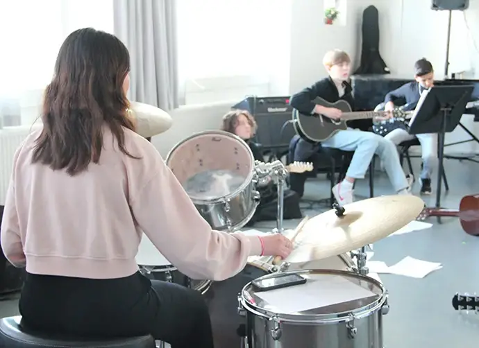 En tjej spelar på ett trumset. I bakgrunden sitter tre ungdomar. En spelar akustisk gitarr och en spelar elbas.