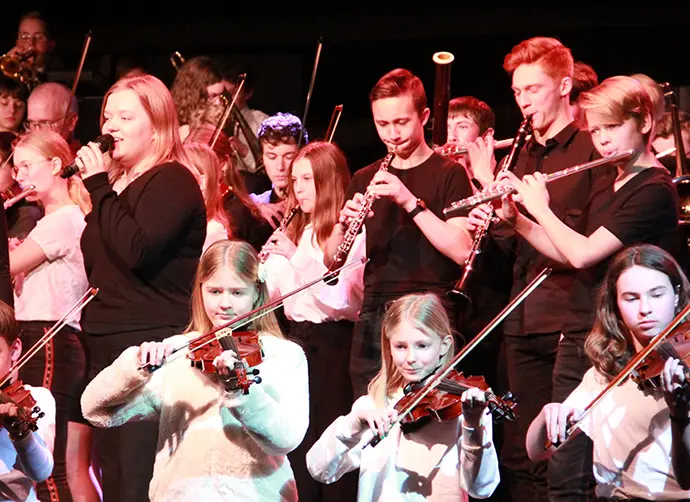 En stor grupp barn och ungdomar spelar tillsammans i orkester. Fioler, klarinetter, tvärflöjt, trumpet, trombon och sång.