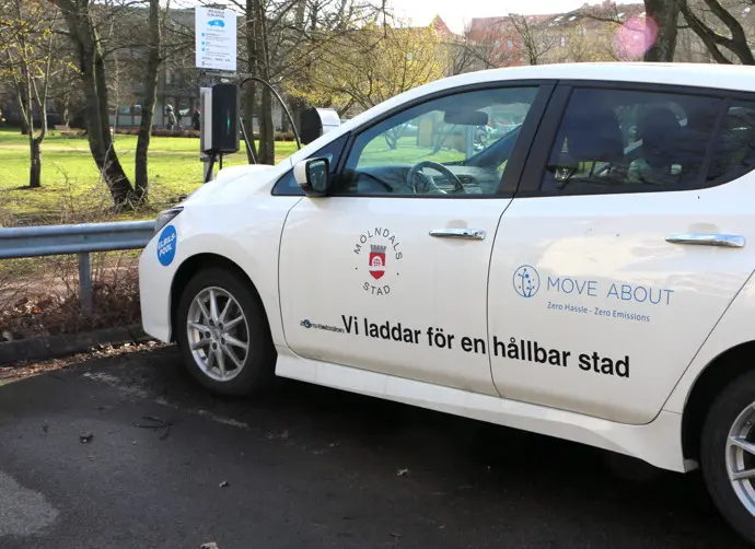 Vit elbil med texten "Vi laddar för en hållbar stad" och logotyper står på en parkering.