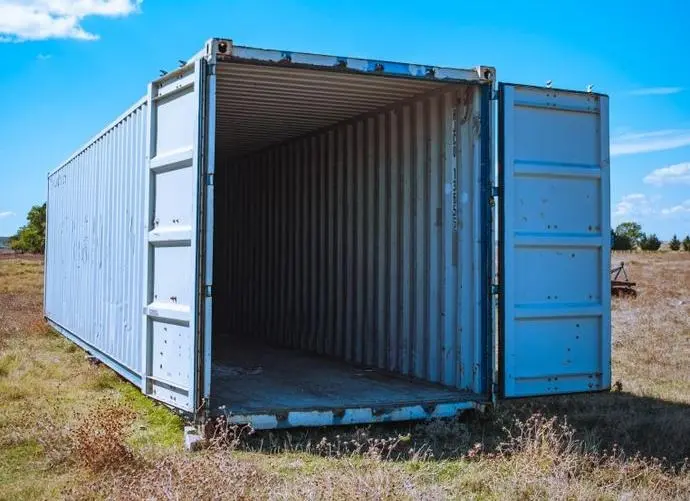 En blå container står öppen på ett fält under en blå himmel. Containern är delvis tom och visar ett mörkt inre.