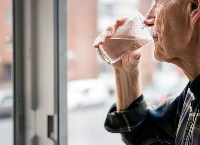 En man står och tittar ut genom ett fönster. I högerhanden håller han ett glas med vatten mot sin mun och dricker.