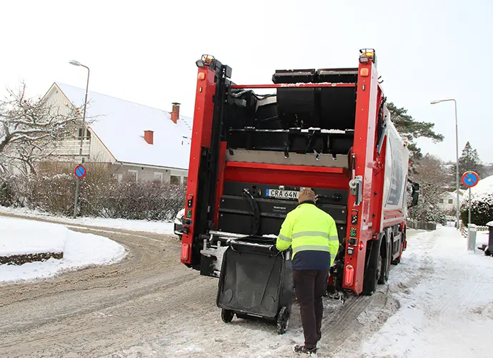 På en gata tävkt med snö står en sopbil. Bakom bilen står en renhållningsarbetare i varselkläder och tömmer ett avfallskärl i bilen.