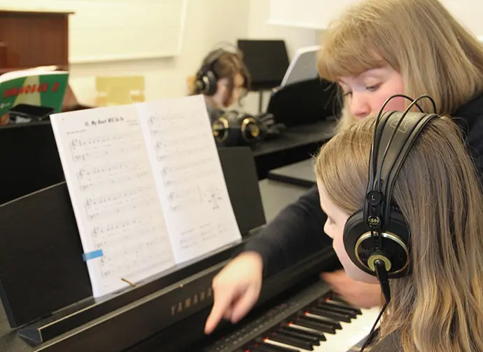 Ett barn sitter vid ett piano med hörlurar på sig. En kvinna står bredvid och pekar på pianotangenterna. På pianot står ett notblad.