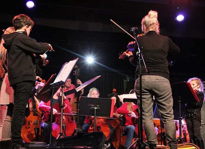 En stråkorkester med barn och lärare spelar tillsammans på en scen. Fioler, cello och kontrabas.