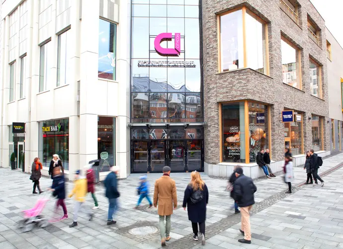 En modern byggnad med affärslokaler och människor som går förbi. På en fasadskylt ovanför entrén står det Mölndals Galleria.