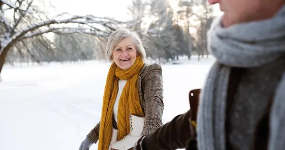 Kvinna med grått hår och gul halsduk med skridskor runt halsen håller hand med en man i snölandskap.