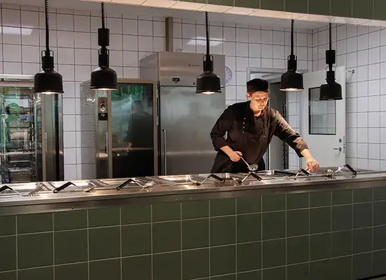 En person i kockkläder arbetar vid en lång rad med tallrikar. Flera mörka lampor hänger från taket i ett restaurangkök.