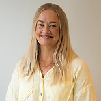 Linda har långt blont hår, ljusgul skjorta och ler mot kameran.