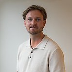 Dominic har på sig en ljus pikétröja, har ljust hår och skägg och ler mot kameran.