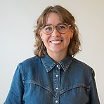 Linda S har kort lockigt hår, glasögon och jeansskjorta och ler mot kameran.