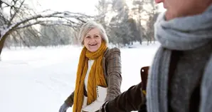Kvinna med grått hår och gul halsduk med skridskor runt halsen håller hand med en man i snölandskap.
